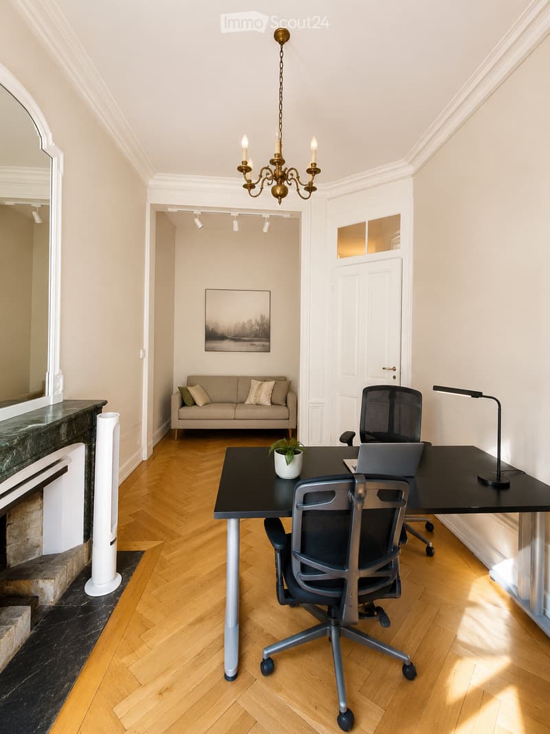 A modern, well-lit room with a wooden floor, a black desk with a laptop, a comfortable chair, a small sofa with pillows, a chandelier, and a potted plant. The room features a fireplace with a mirror above it and a view into another room with a painting on