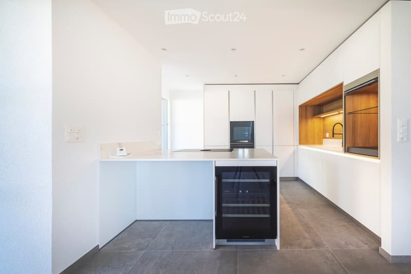 Modern kitchen with white cabinets, a central island, a wine cooler, and a microwave. The space features sleek, minimalist design with integrated lighting and a polished tile floor.
