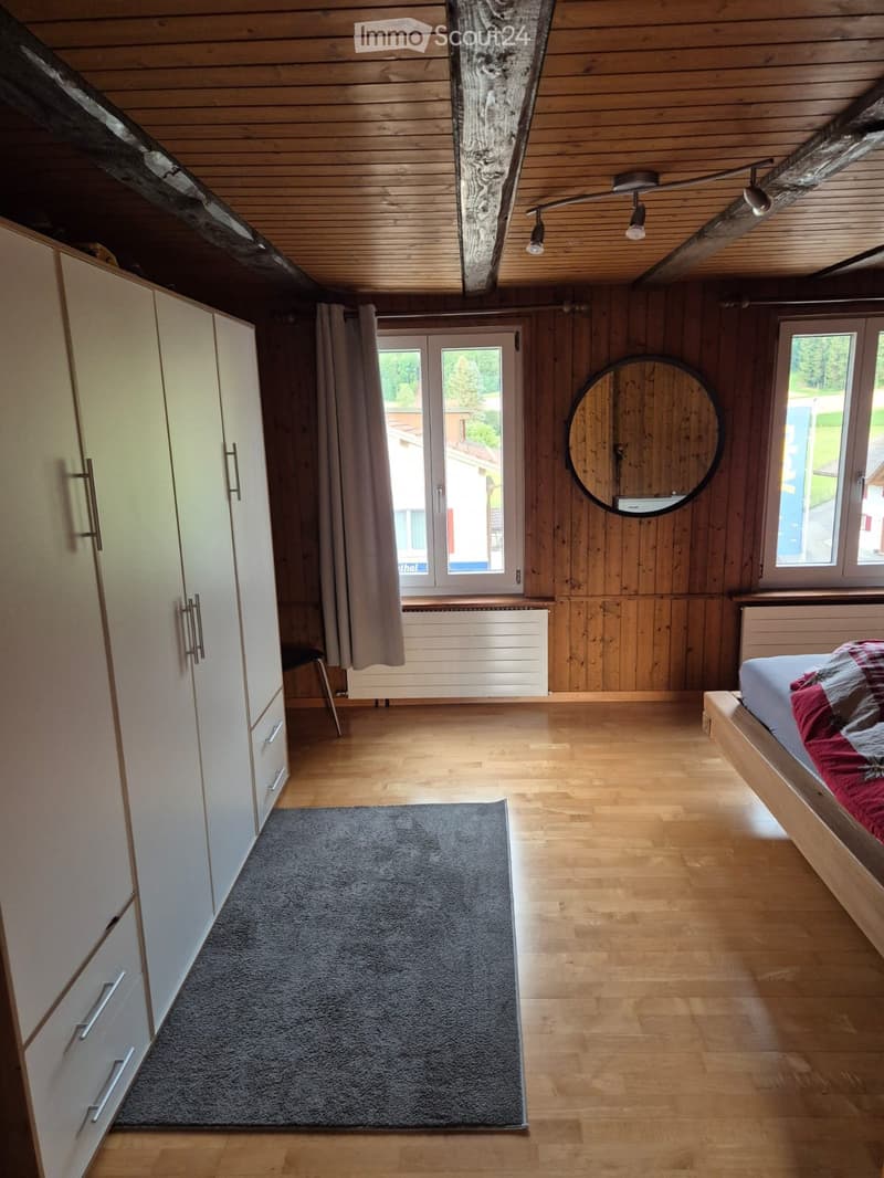 Cozy bedroom with wooden walls and ceiling, featuring a bed with red and grey bedding, a grey rug, a white wardrobe, a round mirror, and a chair near the window.