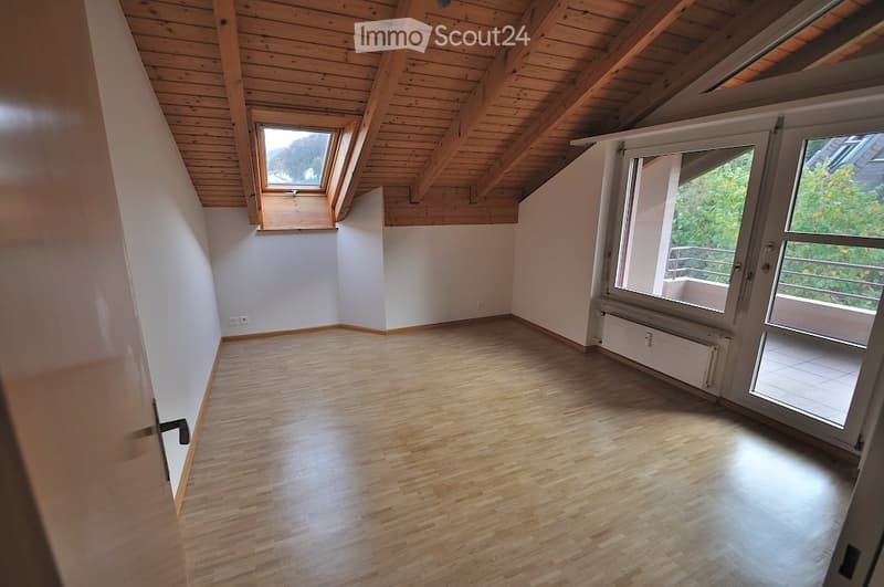 Empty room with wooden flooring, white walls, exposed wooden ceiling beams, and large windows including a skylight and a sliding door leading to a balcony.