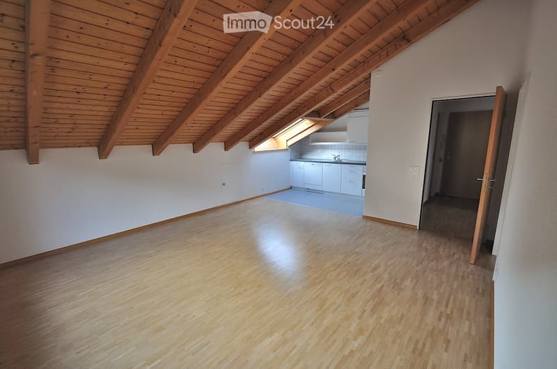 Empty room with wooden flooring and exposed wooden beams on the ceiling. Adjacent kitchen area with white cabinets and a sink is visible through an open doorway.