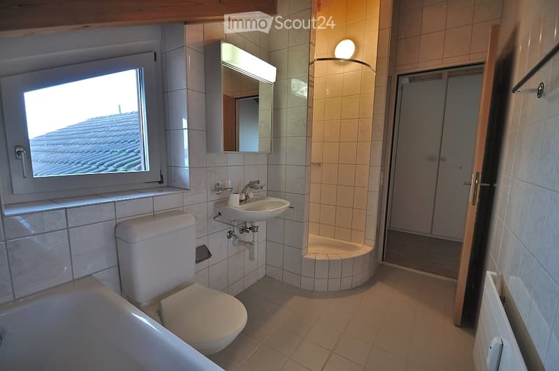 Modern bathroom with a toilet, sink, bathtub, and a corner shower. Features tiled walls and floor, a large window, and a built-in mirror with lighting.
