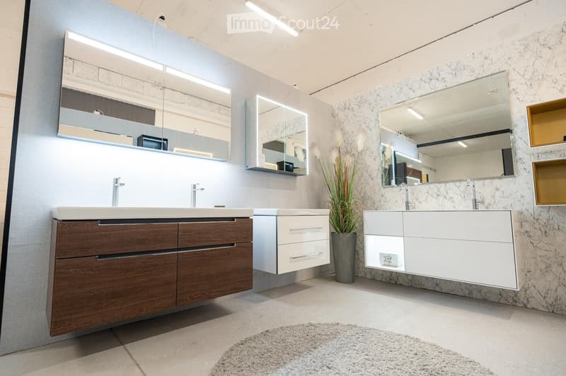 Modern bathroom with two sinks, one with a wooden cabinet and the other with white drawers, large mirrors with built-in lighting, and a decorative plant in a pot.