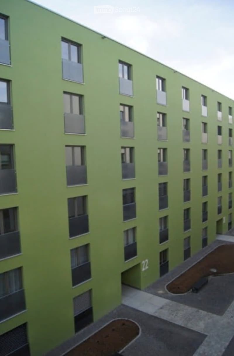 Modern multi-story building with green facade and numerous windows, featuring a numbered entrance and small landscaped areas in front.