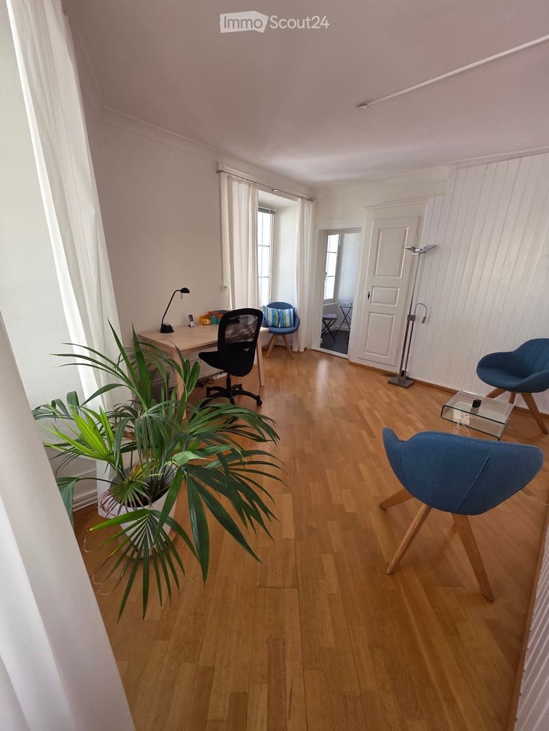 A modern, well-lit room with wooden flooring, white walls, and large windows covered with white curtains. The space includes a desk with a lamp, a potted plant, and several contemporary chairs.