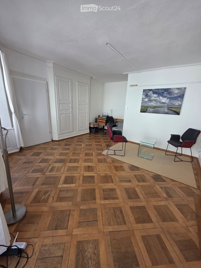 A furnished room with wooden parquet flooring, two chairs, a small glass table, a desk with a laptop, and a large picture on the wall.