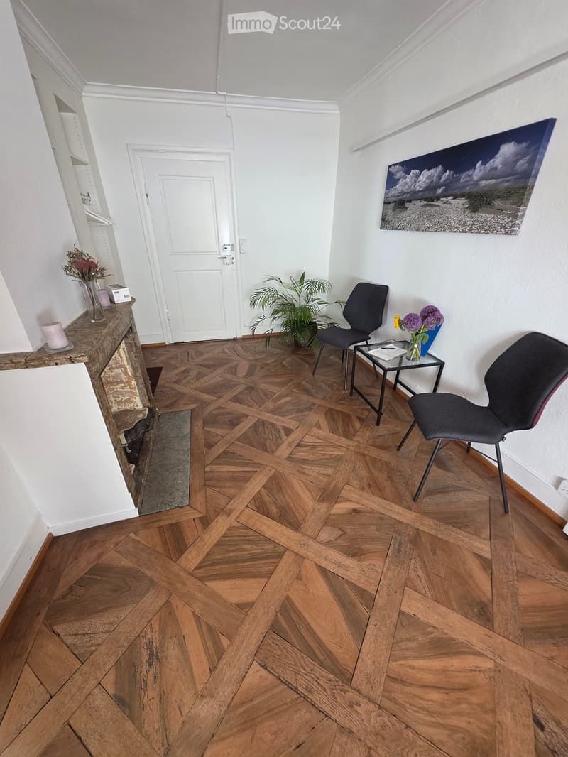 A furnished room with wooden herringbone flooring, white walls, and a fireplace. It features two chairs, a small table with a vase of flowers, a candle, and a potted plant near the door.