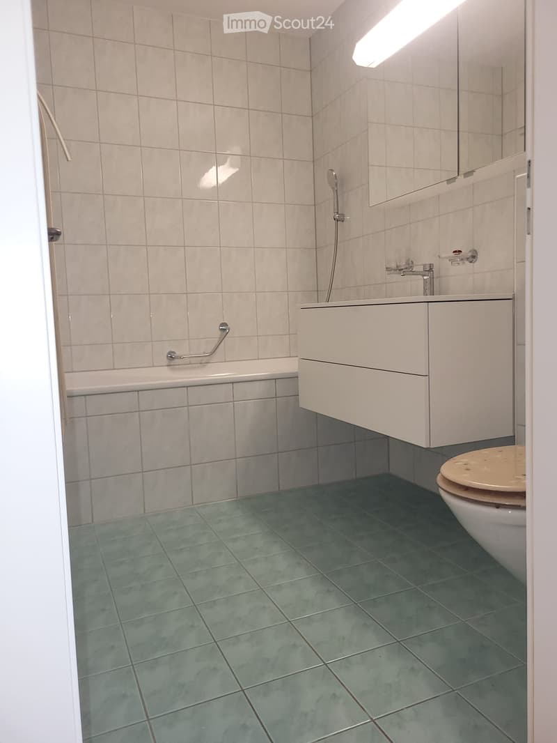 Modern bathroom with a bathtub, toilet, sink, and mirror. The walls are tiled in light beige, and the floor is green tiled.