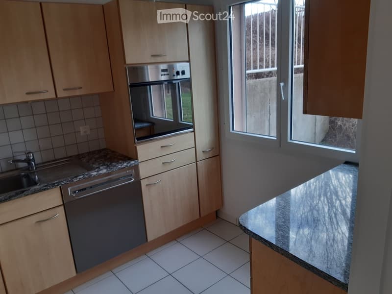 Modern kitchen with light wooden cabinets, granite countertops, stainless steel appliances, and large windows.