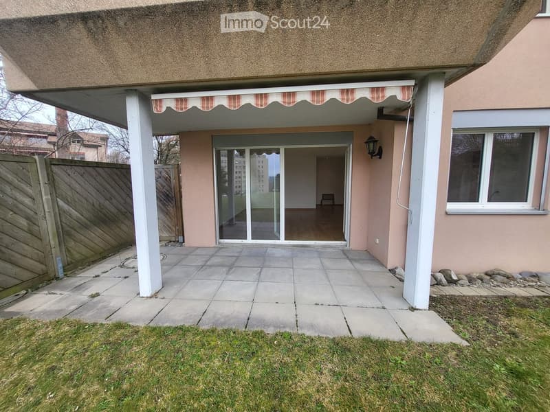 Outdoor patio area with tiled flooring, covered by a striped awning, leading to a glass sliding door into a room. Wooden fence on one side, grassy area in front.