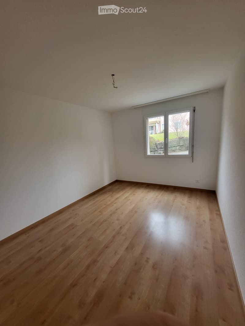 Empty room with light wooden flooring and white walls, featuring a large window with a view of the outside.