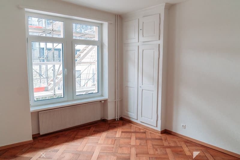 Empty room, large window, white walls, wooden parquet flooring, white built-in wardrobes, under-window radiator, electrical outlet.