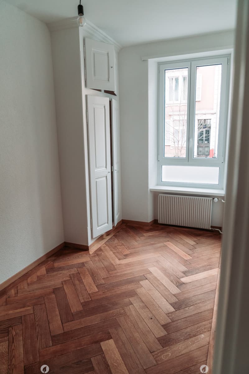 Empty room with wooden herringbone floor, white walls, double doors wardrobe, and window with radiator.