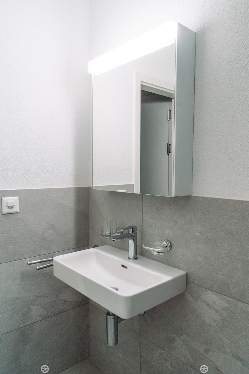 bathroom with white sink, gray tiled walls, modern mirror with integrated light