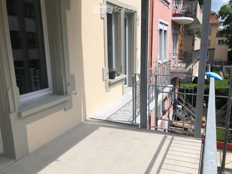 Balcony with metal railing, concrete floor, and view of neighboring buildings and greenery.