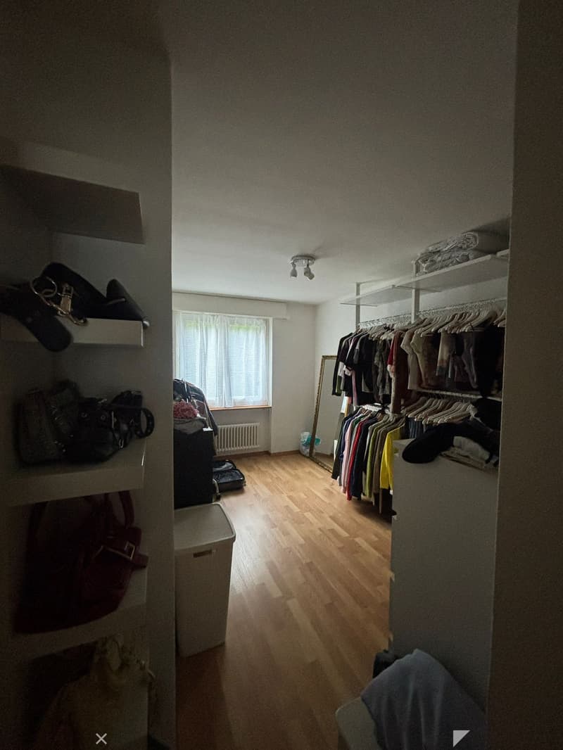 A well-lit, organized walk-in closet with wooden flooring, a large wardrobe filled with hanging clothes, a mirror, and various storage items including bags and a suitcase.