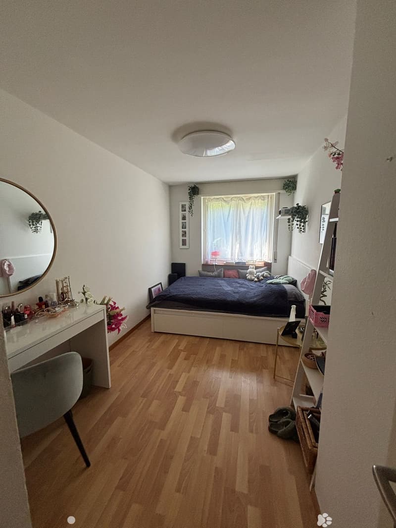 Cozy bedroom with wooden flooring, a bed with dark bedding, a vanity with a mirror, a chair, and a small table with decorative items.
