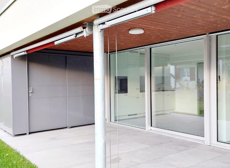 Modern outdoor patio with large sliding glass doors leading to a kitchen area, covered by a retractable awning, and a grey garage door.