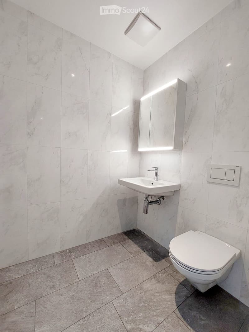 Modern bathroom with white marble tiles, a white sink, a toilet, and a mirrored cabinet with lighting.