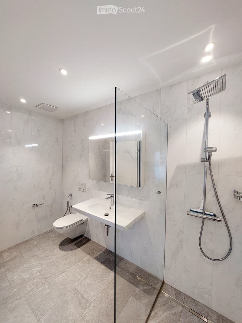 Modern bathroom with white marble tiles, a glass-enclosed shower, a toilet, a sink with a mirror, and a modern showerhead.