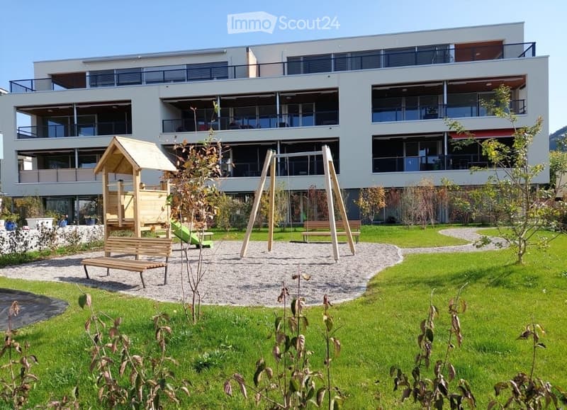 Modern multi-story building with balconies, surrounded by a well-maintained garden area featuring a playground with swings, benches, and a small wooden playhouse.