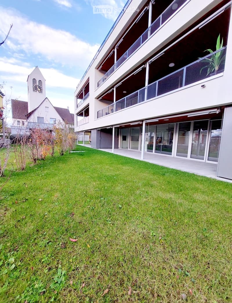 A well-maintained lawn in front of a modern multi-story building with balconies and large windows. The building has a contemporary design with clean lines and glass panels.