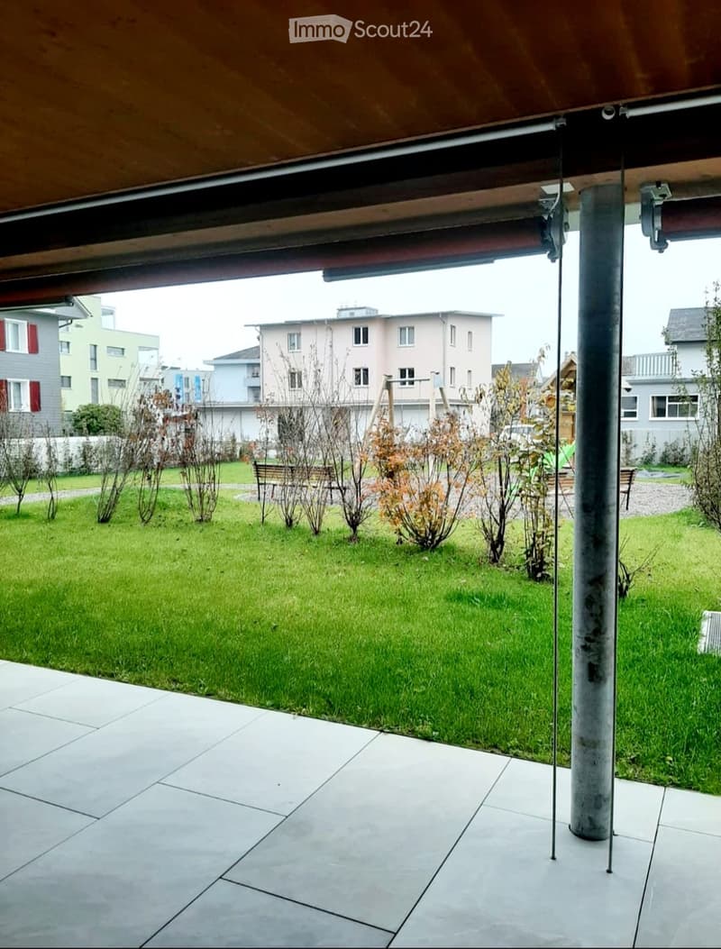 A modern outdoor patio with a tiled floor, a grassy lawn, and surrounding residential buildings in the background.