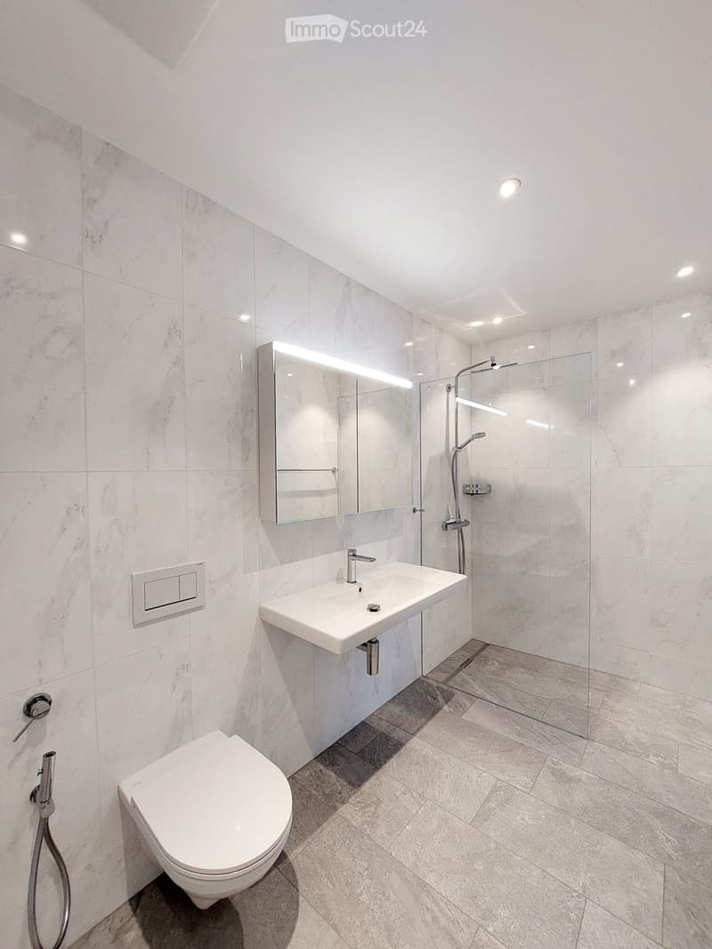 Modern bathroom with white marble tiles, a toilet, a sink with a modern faucet, a large mirror with LED lighting, and a glass-enclosed shower area.