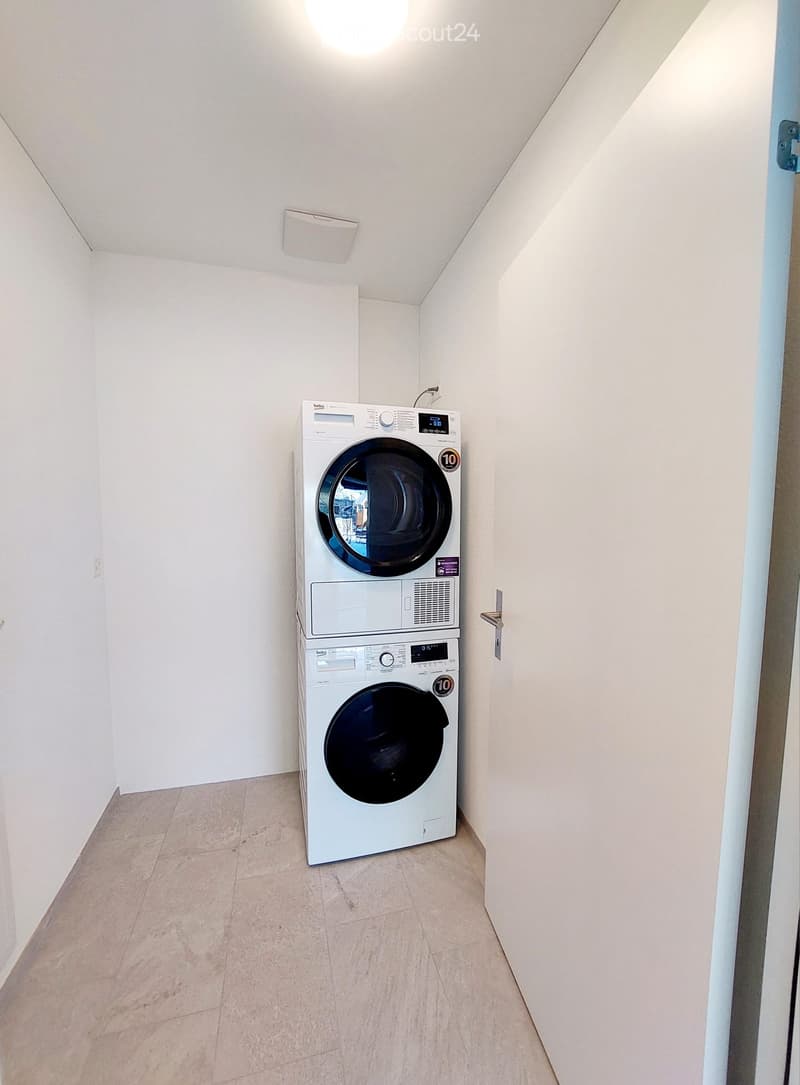 A small, well-lit room with a stacked washer and dryer unit against one wall, light-colored tiled floor, and white walls.