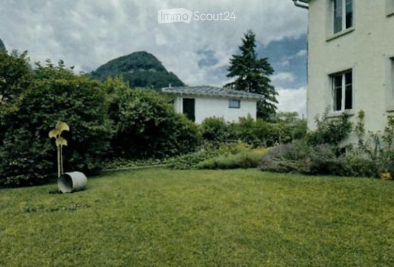 Well-maintained garden with green grass, bushes, and trees, featuring a scenic mountain view in the background.