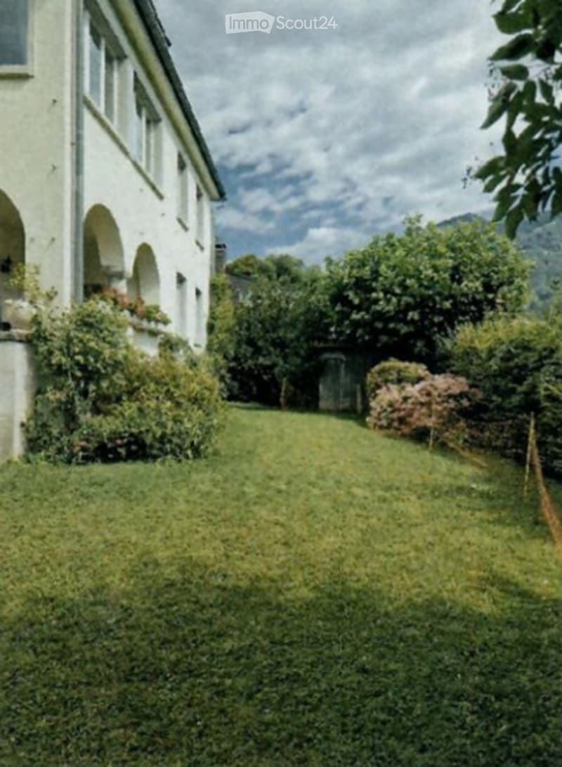 A well-maintained garden area with a lush green lawn, surrounded by a white building with arched windows and greenery. The background features trees and a scenic view of mountains under a partly cloudy sky.