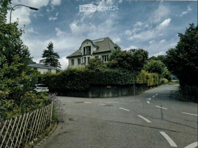 A large, multi-story house with a pitched roof and multiple windows, surrounded by greenery and a low hedge. The house is partially obscured by trees and a wooden fence along the street.
