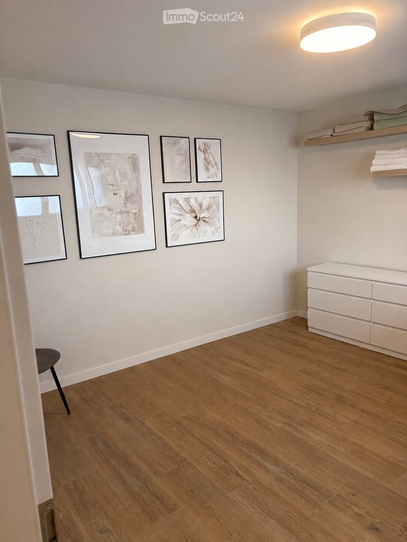 Empty room with wooden flooring, white walls, and modern decor including framed artwork and a small cabinet with shelves.