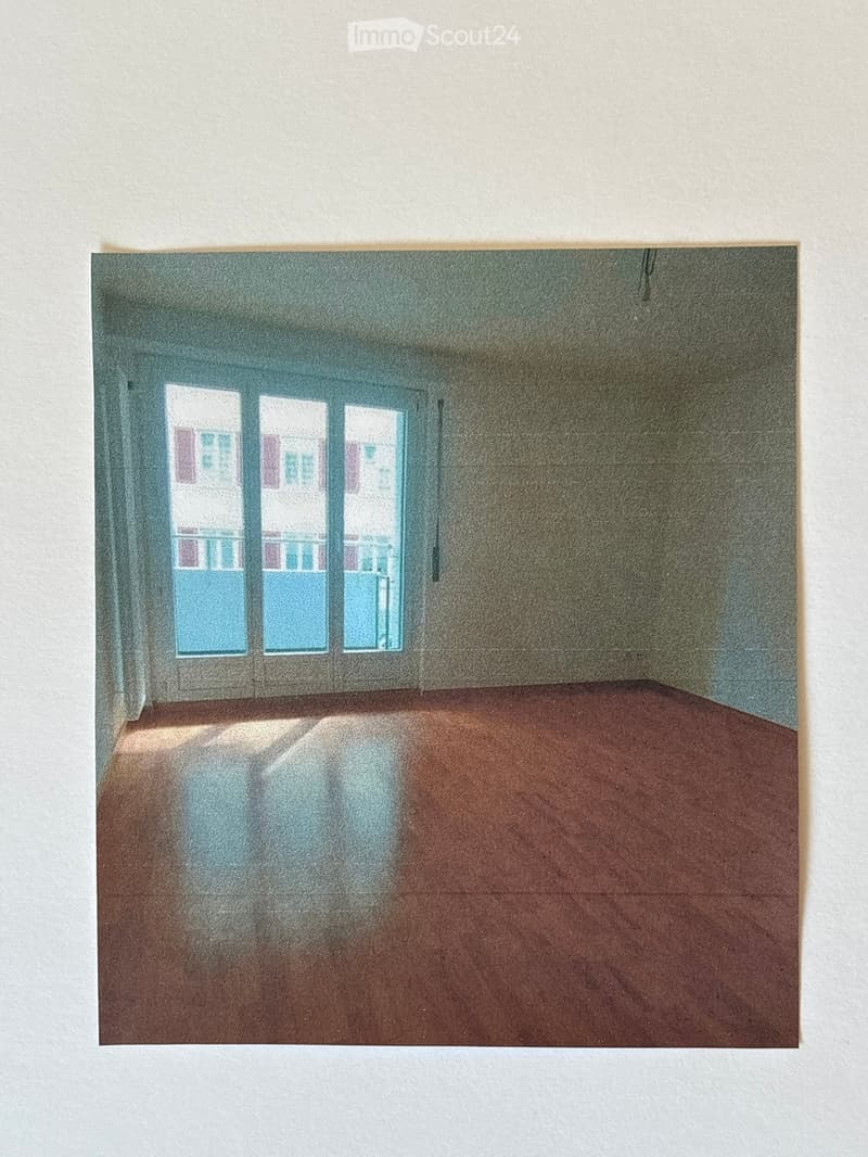 Empty room with wooden floor and large windows showing a view of buildings outside. No furniture or equipment present.