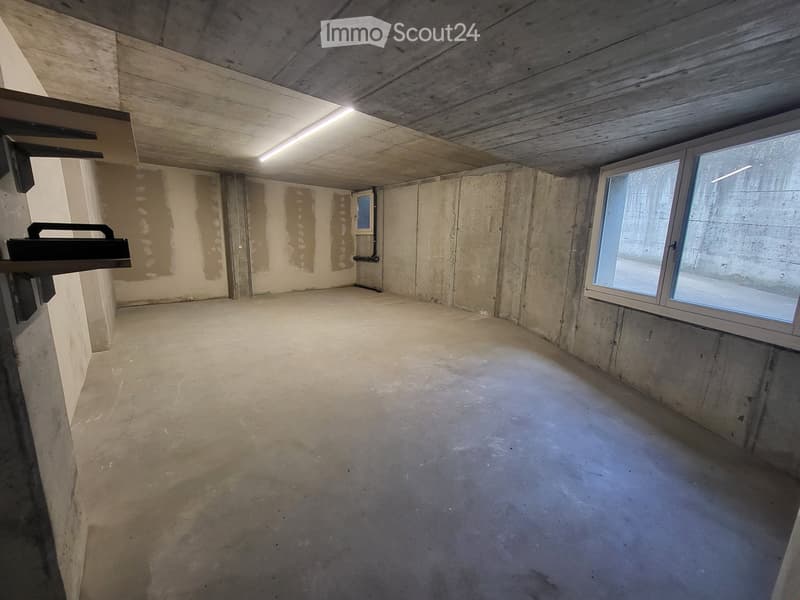 Empty room with concrete walls and floor, wooden ceiling, and a single window letting in natural light.