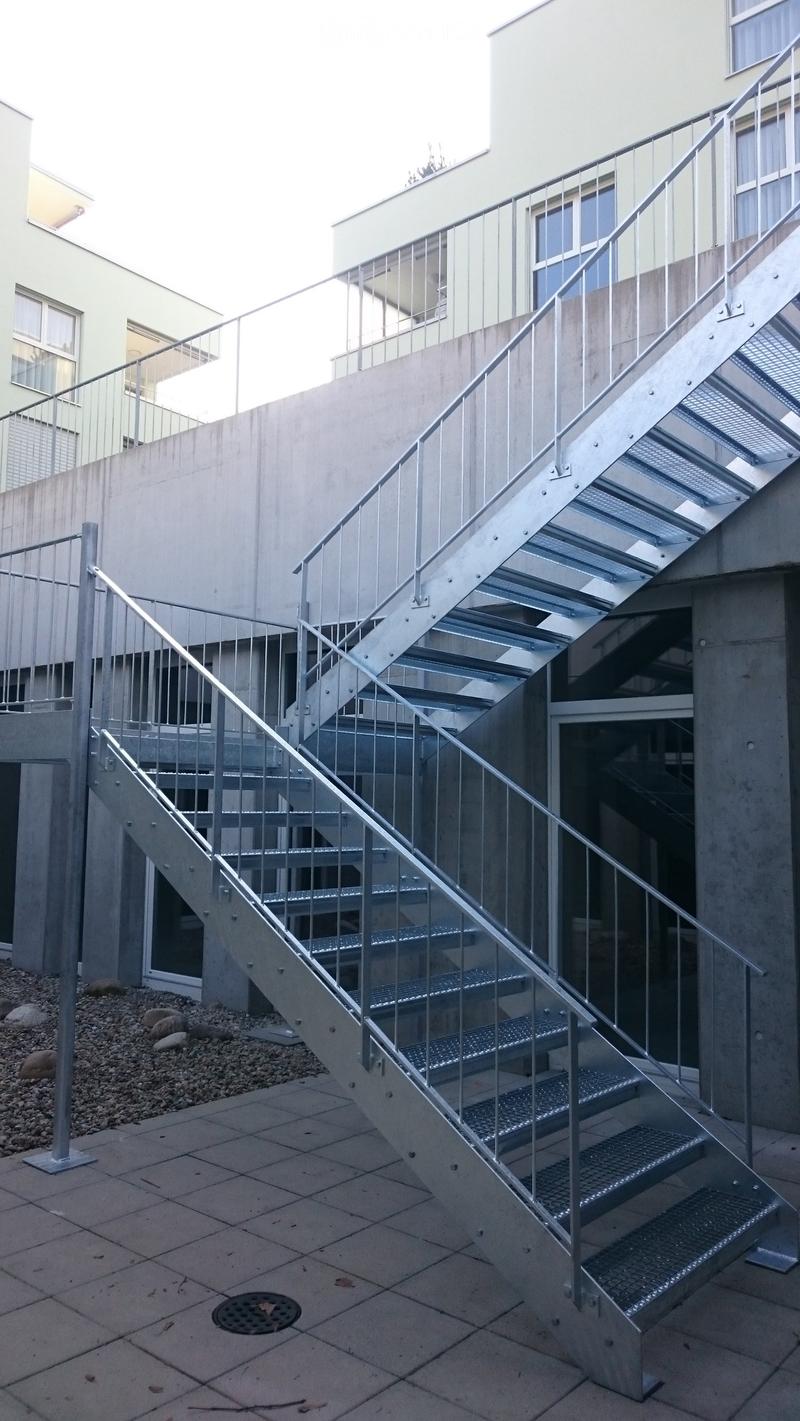 Metal staircase with handrails leading up to a raised ground floor area, surrounded by concrete walls and a tiled ground.