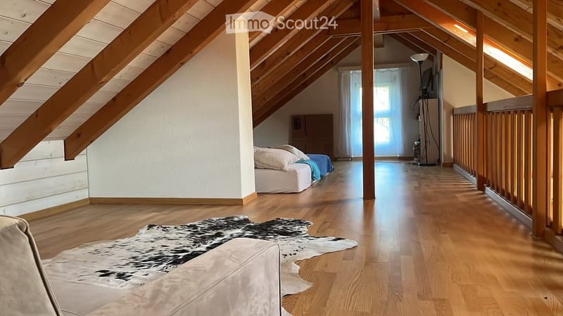 Spacious attic room with wooden beams, hardwood floors, a bed, a sofa, a large cowhide rug, and large windows with curtains.