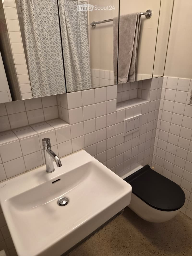 Modern bathroom with white tiles, a white sink with a chrome faucet, a toilet with a black seat, a mirror with patterned curtains, and a towel on a rack.