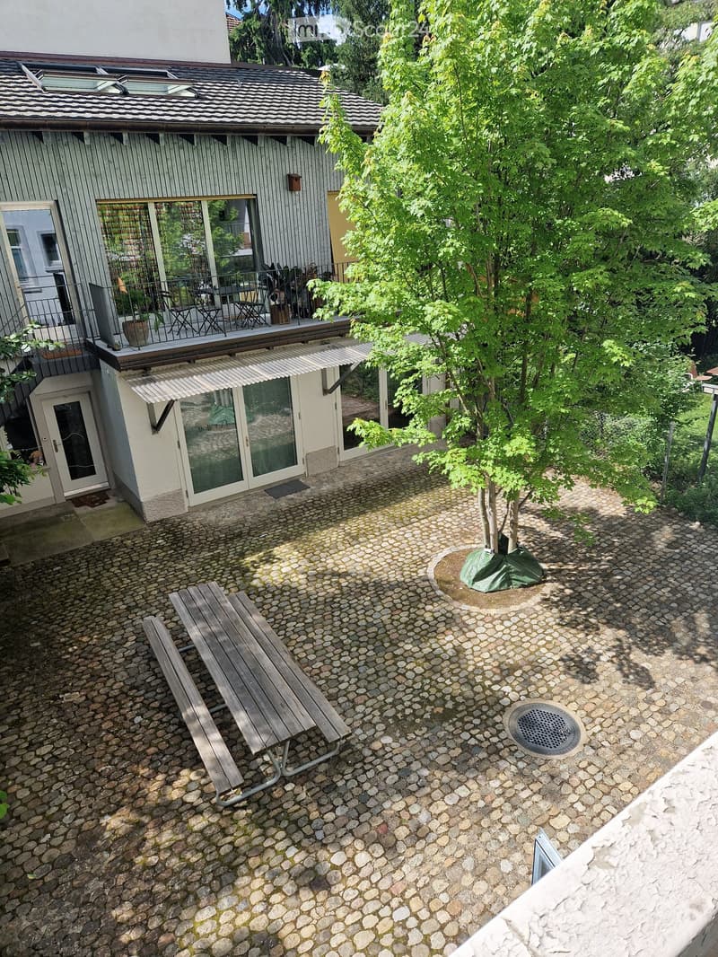 Pebbled outdoor area with a wooden bench, small tree, and a covered entrance to a building with a balcony and outdoor seating area.