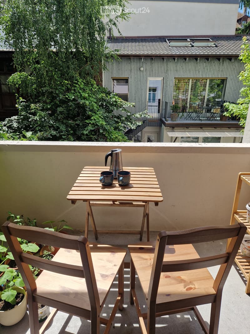 Balcony with wooden table, two chairs, coffee pot, cups, and potted plants.