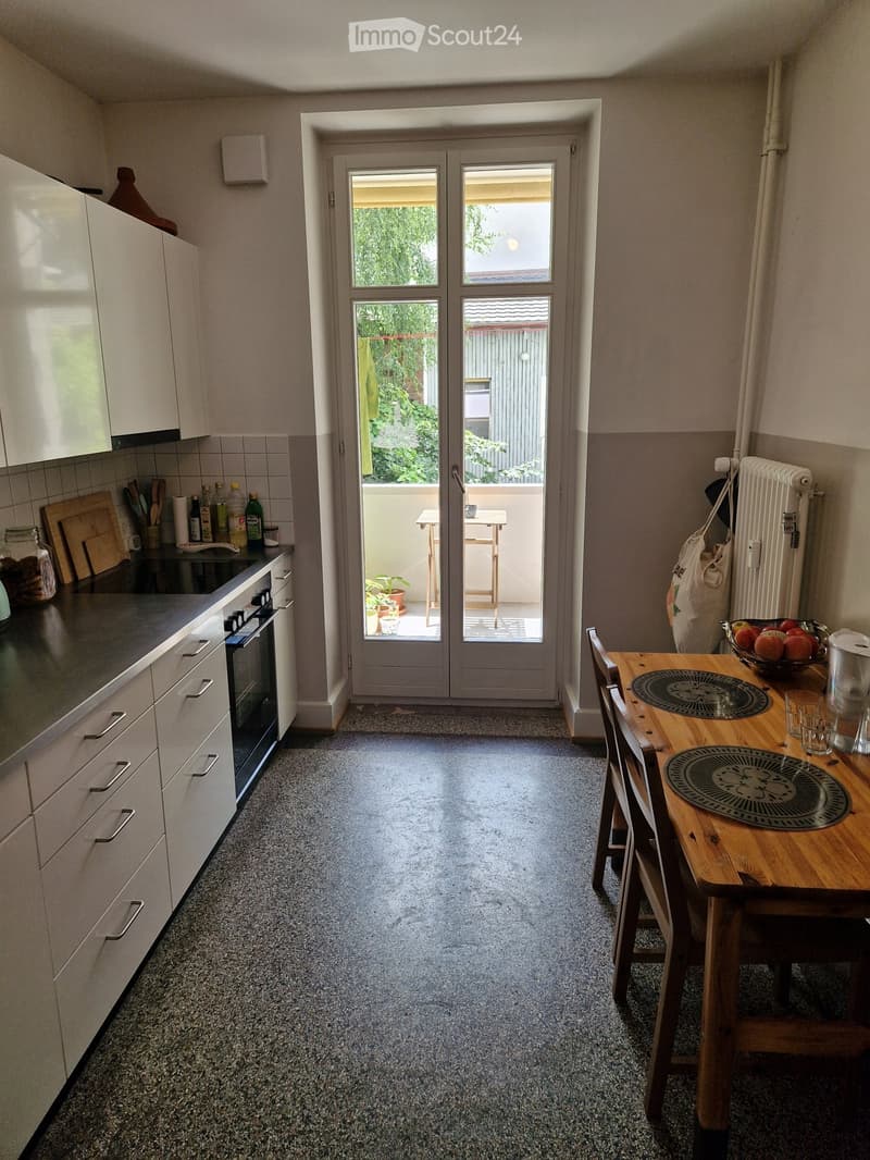 Modern kitchen with white cabinets, grey countertop, and a dining area with a wooden table and chairs. A sliding glass door leads to a small balcony with outdoor furniture.