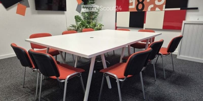 A modern meeting room with a white rectangular table surrounded by red and black chairs, a potted plant, and colorful wall art.