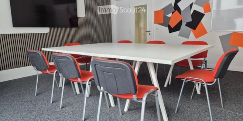 A modern, minimalist meeting or break room with a white rectangular table surrounded by colorful chairs, a large flat-screen TV mounted on the wall, and a decorative geometric wall design.