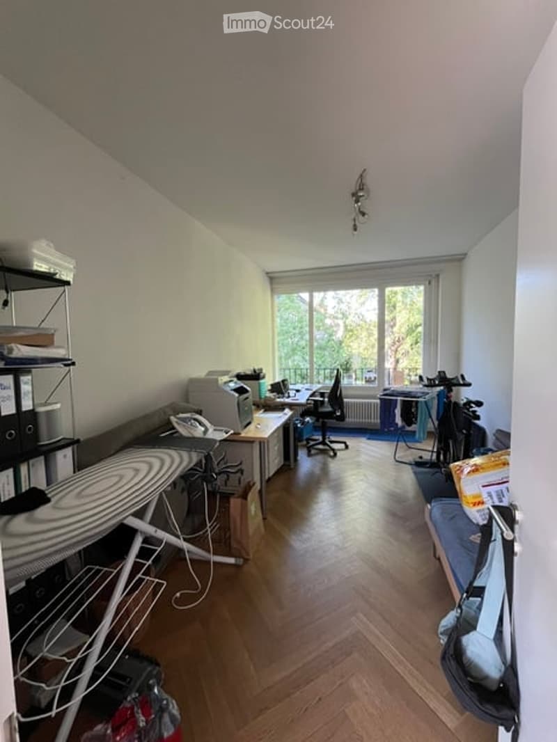 A multi-functional room with a wooden floor, large windows offering a view, a desk with a chair, a printer, an ironing board, shelves, a laptop, a radiator, a bicycle, and a small sofa.