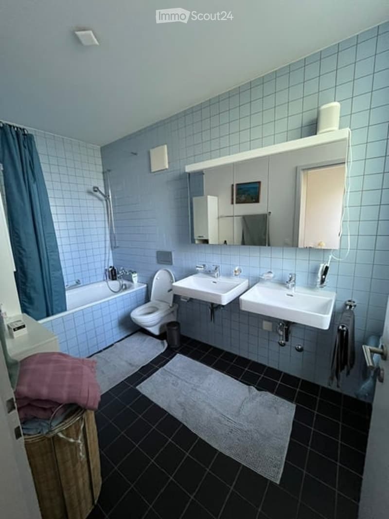 Bathroom with blue tiled walls, black tiled floor, bathtub, toilet, two sinks, mirror, and towels.