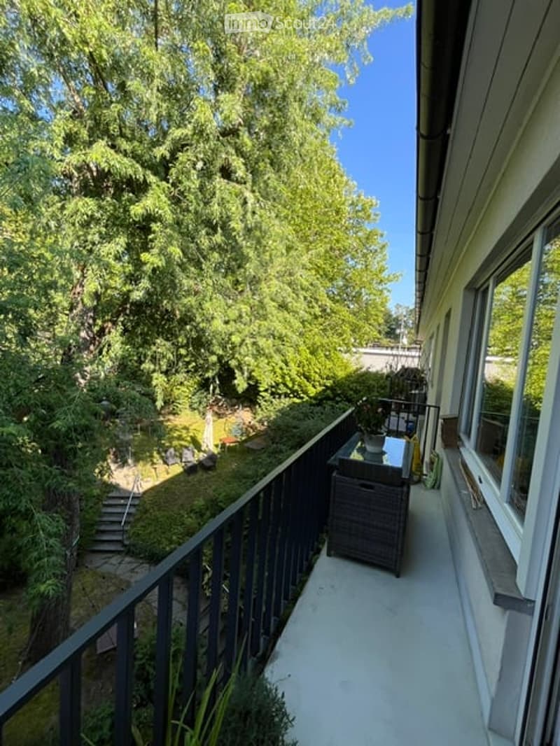 Balcony with a view of a lush garden, featuring a small table and chair, railing, and steps leading down to the garden.