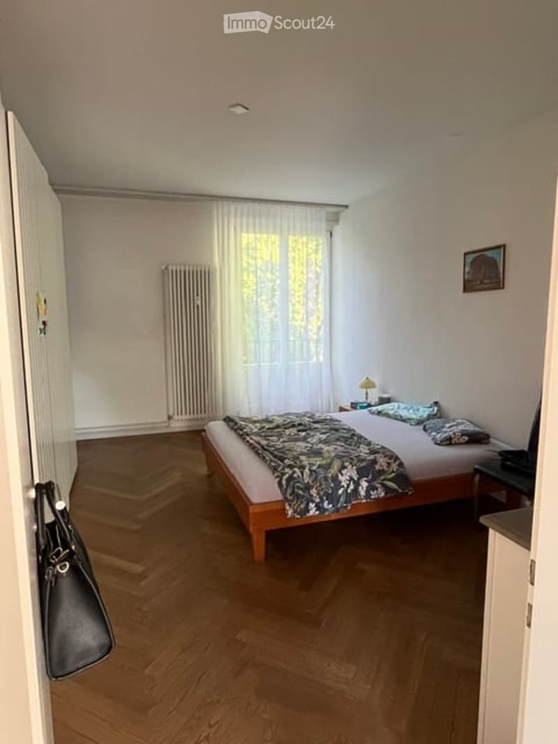 Cozy bedroom with wooden flooring, a double bed with a patterned duvet, a nightstand with a lamp, a radiator, and a picture frame on the wall.