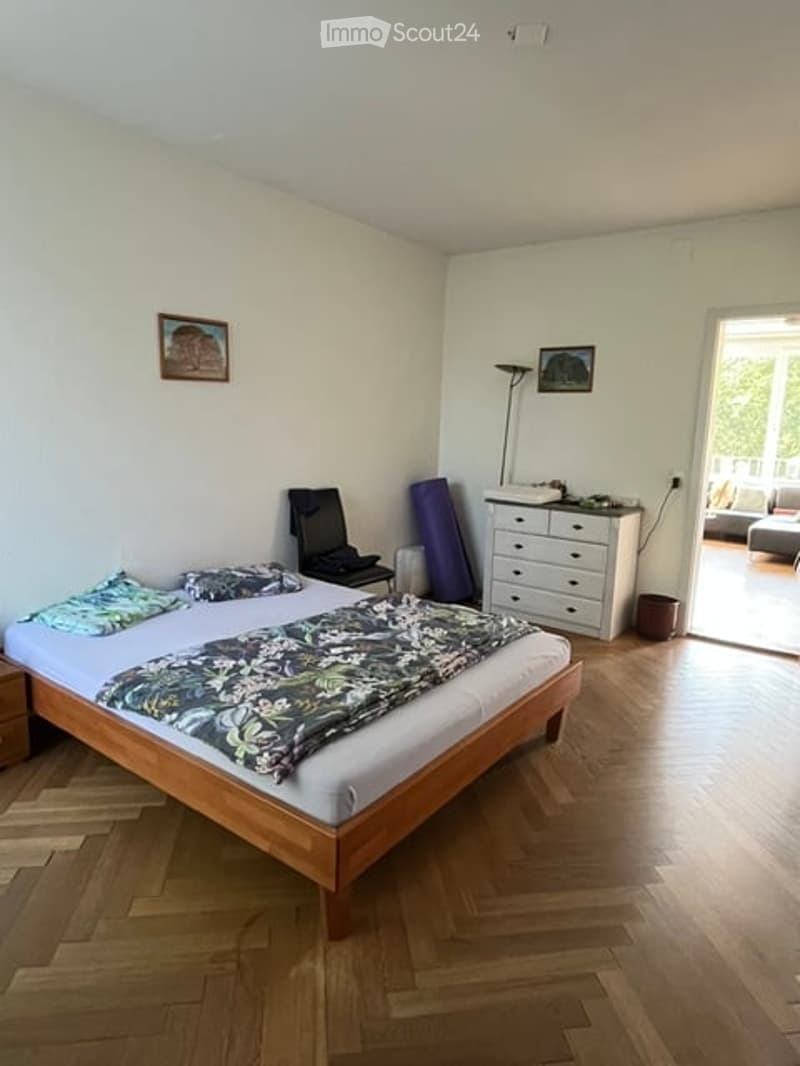 Bright bedroom with wooden flooring, a neatly made bed with patterned bedding, a nightstand, a chair, a dresser, and a floor lamp. A picture frame is on the wall, and the room has large windows letting in natural light.