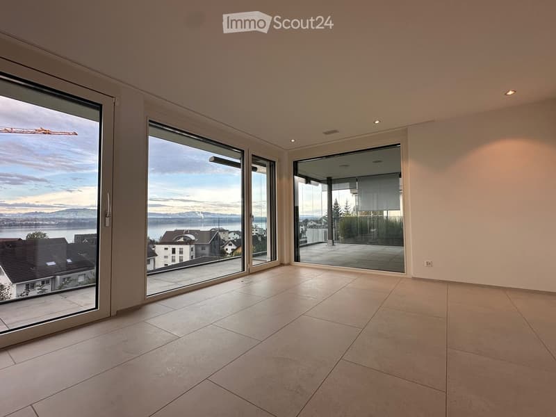 Empty room with large glass sliding doors leading to a balcony, tiled floor, and a scenic view of a lake and town.