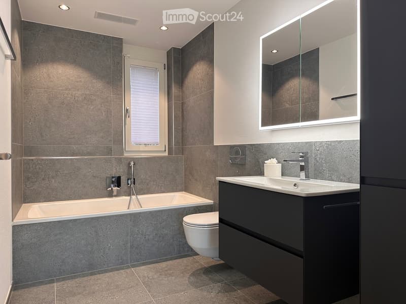 Modern bathroom with grey tiled walls, a bathtub, toilet, sink, and a large illuminated mirror above the sink.